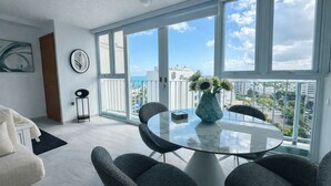 Een moderne woonkamer met een glazen tafel, grijze stoelen, een vaas met bloemen, een klok aan de muur en een uitzicht op de stad door grote ramen.