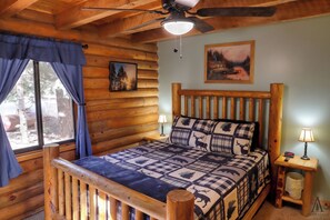 Une chambre dans un chalet en rondins, avec un lit, un ventilateur de plafond, une fenêtre avec des rideaux, une table de chevet avec une lampe et un tableau accroché au mur.