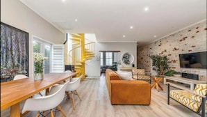 A modern living room with a wooden dining table, white chairs, a brown sofa, and a TV. - 3 Bedroom Home in Gardens Cape Town (Cape Town)