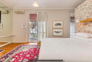 Un dormitorio con cama, alfombra estampada, pared con textura de ladrillo y puerta que da a un espacio exterior.