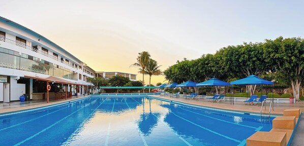 A swimming pool with lounge chairs and umbrellas, a bar area, and a building with multiple floors and balconies.