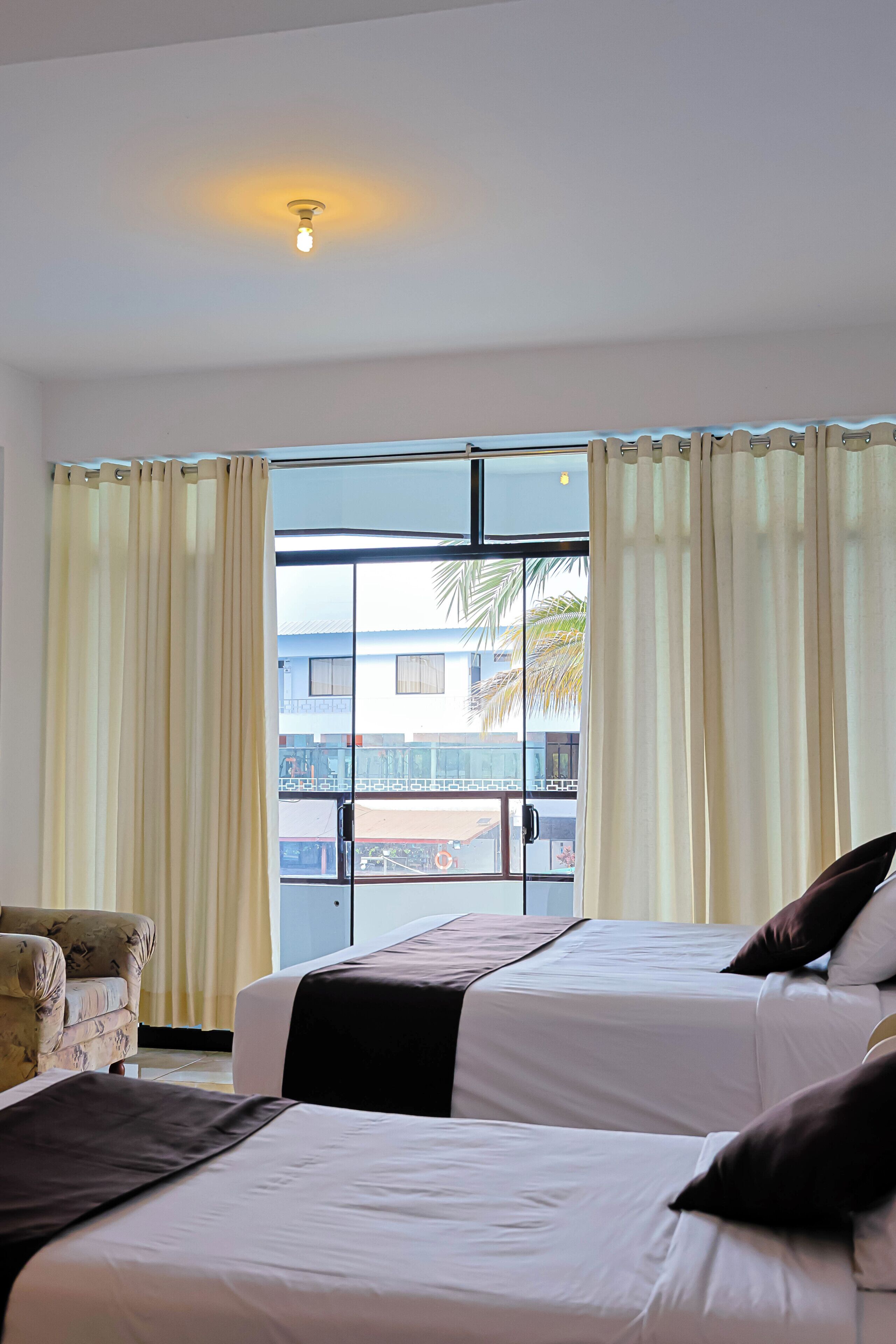 A hotel room with two beds, a sofa, a window with curtains, and a view of a building and palm trees outside.