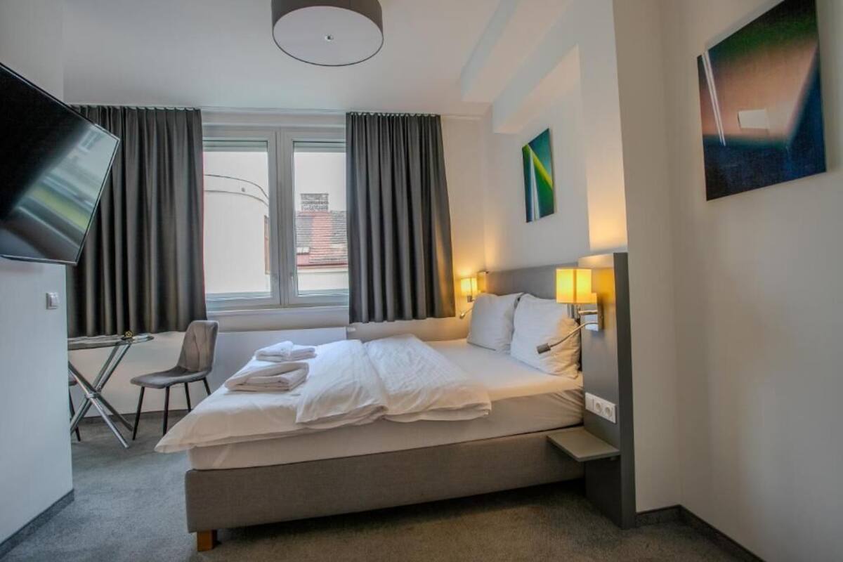 A hotel room with a bed, bedside table, lamp, TV, chair, and a window with curtains.