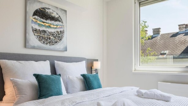A neatly made bed with white linens and teal pillows, a wall-mounted artwork, and a window with a view of buildings. - Cube Apartments by Wiener Stadthalle (Vienna)