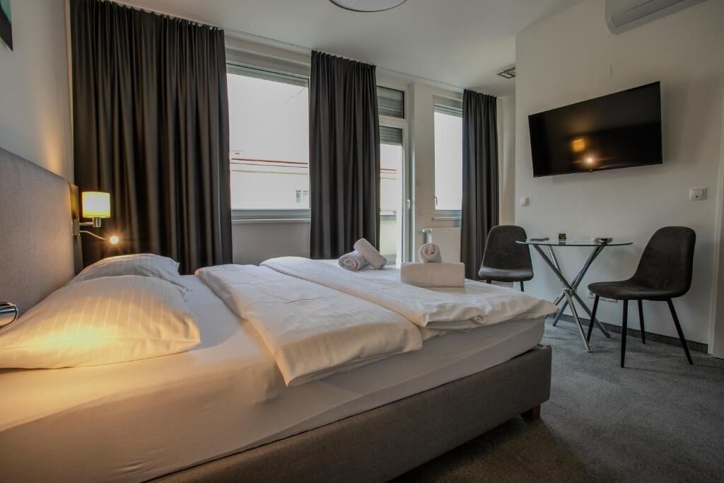 Et hotelværelse med seng, fjernsyn, skrivebord med to stole og store vinduer med gardiner.