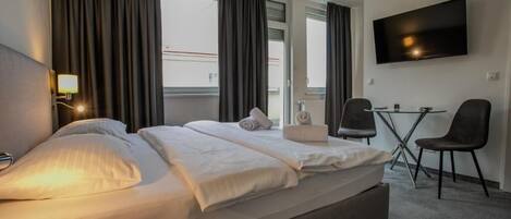 Et hotelværelse med seng, fjernsyn, skrivebord med to stole og store vinduer med gardiner.