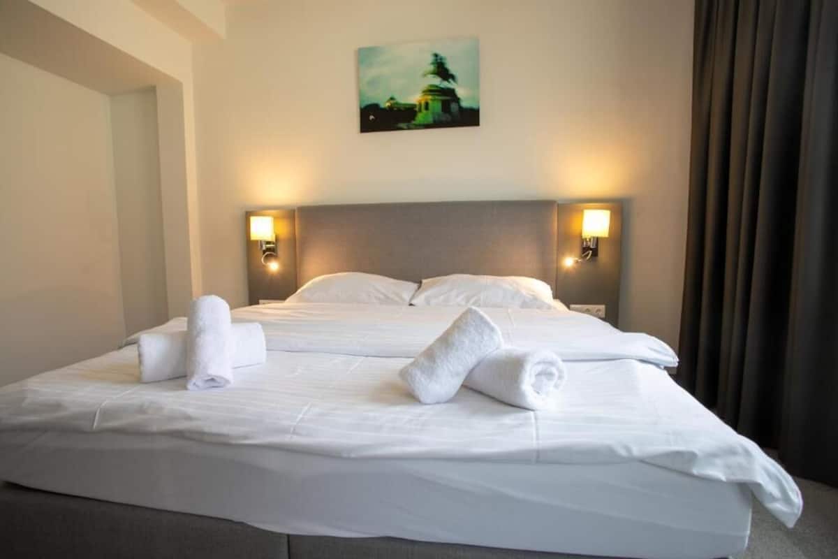 A neatly made bed with white linens and towels, two wall-mounted lamps, and a painting on the wall.