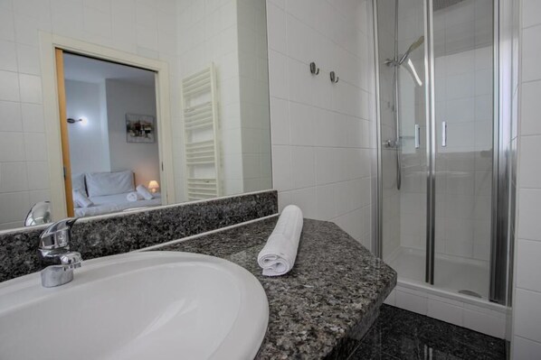 Un baño con lavamanos blanco, espejo, toalla y ducha con mampara de vidrio.