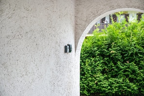 Eine strukturierte Wand mit einer Überwachungskamera und einem Durchgang zu einer grünen Hecke.