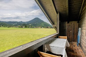 Un interior de madera con una mesa y sillas, vista a un campo verde y casas a lo lejos, y una montaña al fondo.