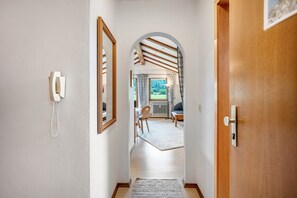 Ein Korridor mit einer Holztür, einem Telefon und Blick in einen Raum mit einem Sessel und einem Tisch.