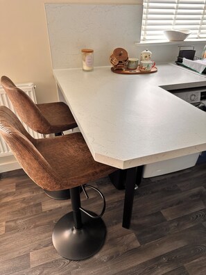A kitchen with a white countertop, two brown upholstered chairs with black bases, a white cabinet, and a window with blinds.