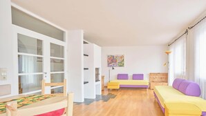 Una habitación con suelo de madera, una cama con ropa de cama amarilla y púrpura, un banco de madera, una mesita con mantel colorido y una ventana con cortinas.