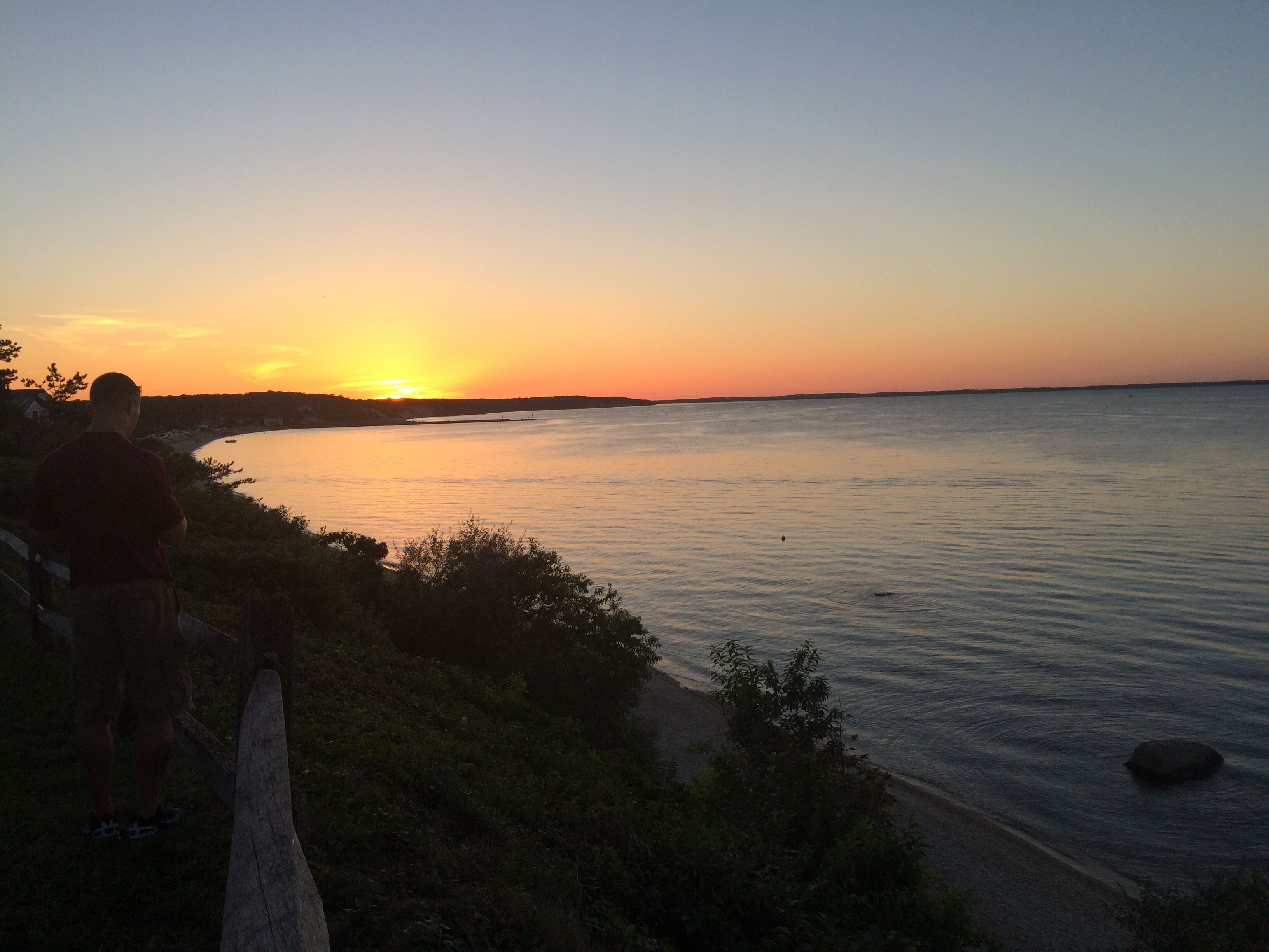 A sunset over a calm body of water with a person standing on a grassy area.