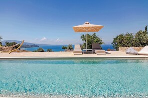 Una piscina cristalina con sillas de descanso y una sombrilla, con vistas a un paisaje costero.