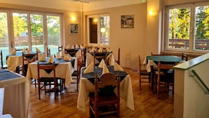 A restaurant with multiple tables and chairs, large windows with views of trees, and a wooden floor. - Garnì Pirata (Cavareno)