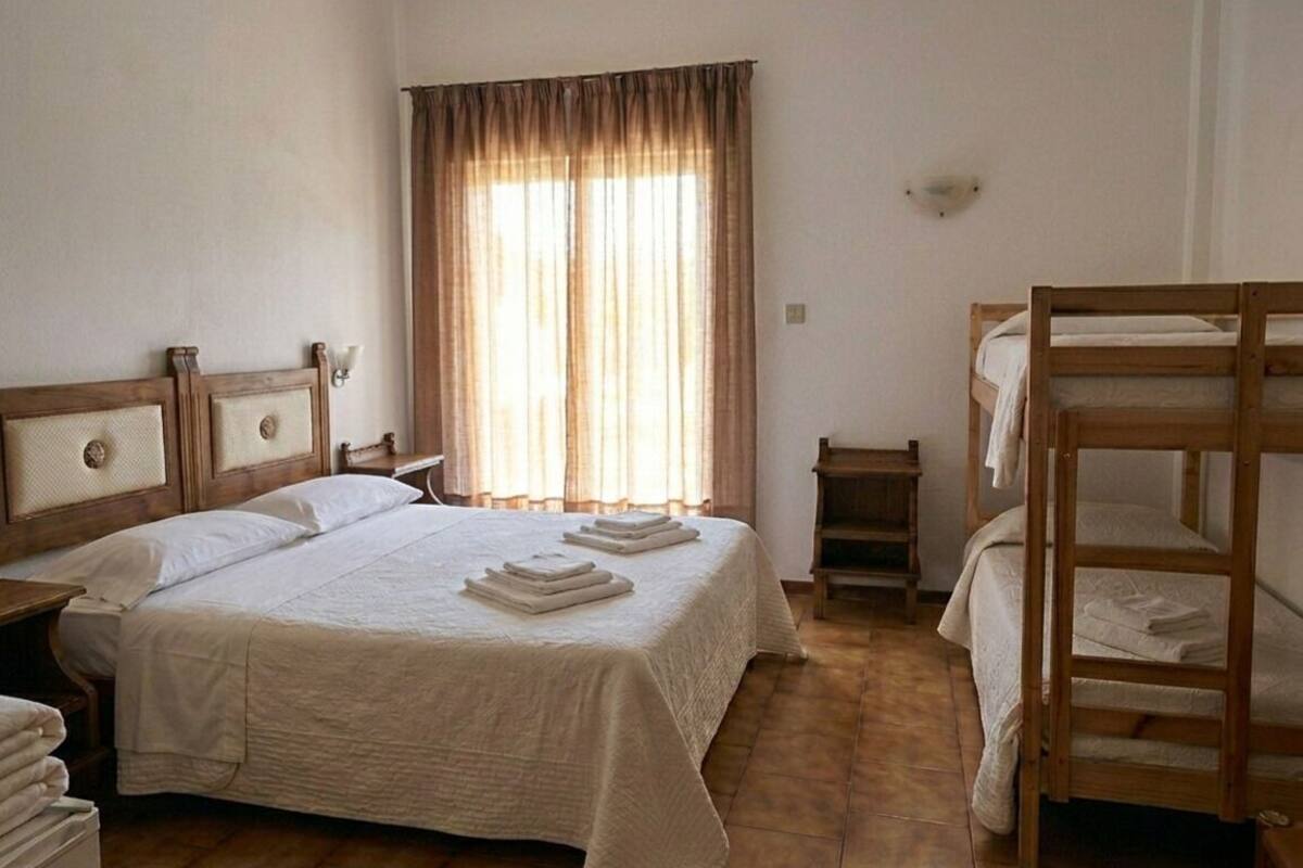 Un dormitorio con una cama grande, una cama más pequeña, una cama nido de madera, una mesita de noche, una lámpara y una ventana con cortinas.