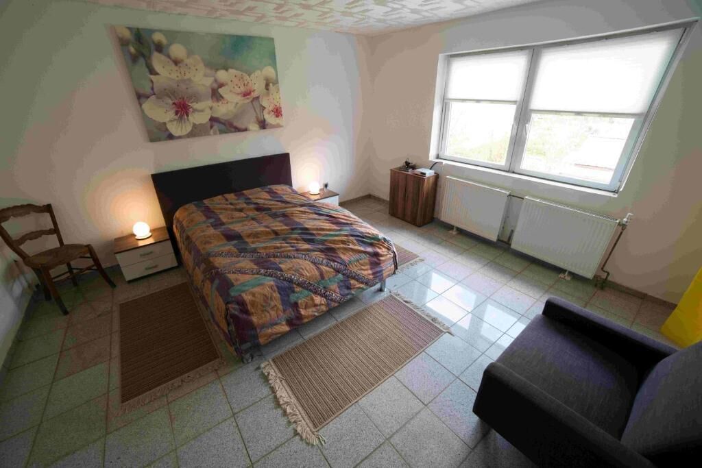 Um quarto com cama, uma cadeira, mesa de cabeceira, radiador e janela.