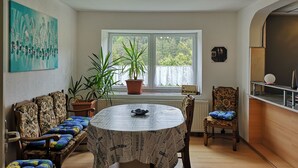Ein Zimmer mit rundem Tisch, einer Couch, einem Sessel, einer Zimmerpflanze, einem Heizkörper und einem Fenster mit Blick auf Grünflächen.