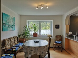 Ein Zimmer mit rundem Tisch, einer Couch, einem Sessel, einer Zimmerpflanze, einem Heizkörper und einem Fenster mit Blick auf Grünflächen.