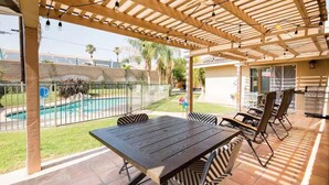 Eine Terrasse im Freien mit Holztisch und -stühlen, einem Pool im Hintergrund und einem überdachten Bereich mit einer Pergola.