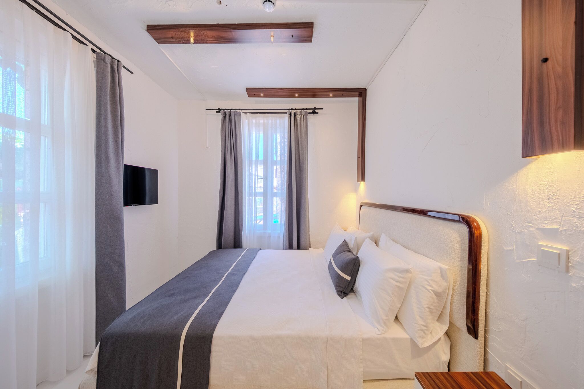 A hotel room with a bed, curtains, a mounted TV, and a wooden headboard.