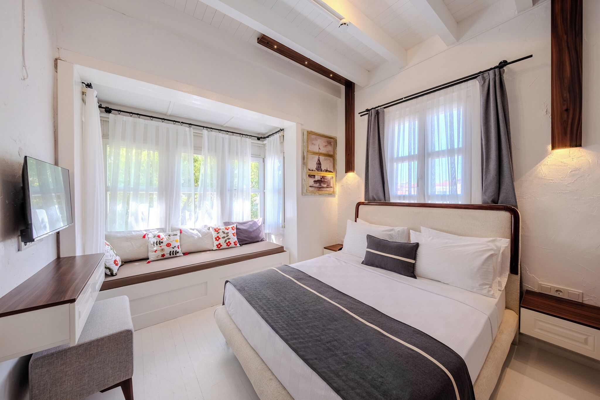 A bedroom with a bed, a desk, a chair, a window with curtains, and a wall-mounted TV.