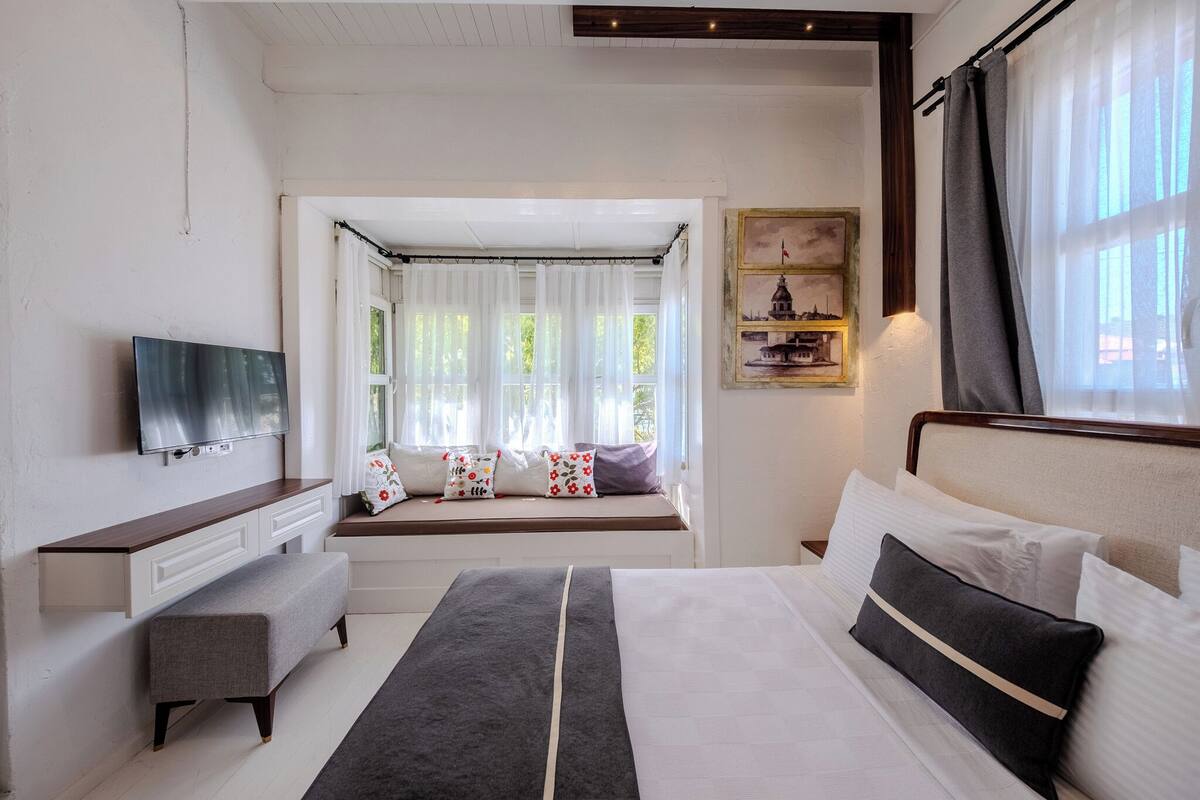 A hotel room with a bed, a bench, a TV, a window with curtains, and a painting on the wall.