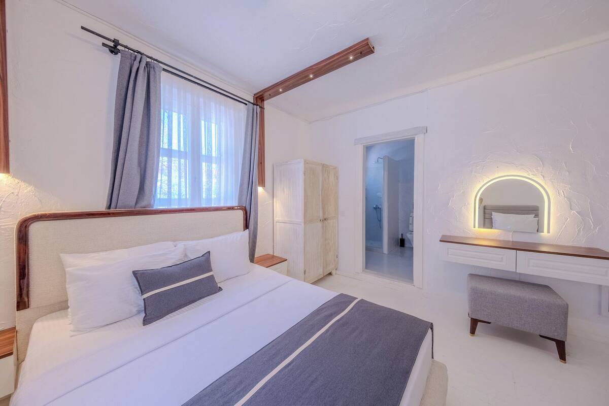 A hotel room with a bed, a bedside table, a wardrobe, a vanity with a mirror, and a door leading to a bathroom.