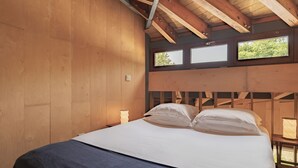 Een houten kamer met een bed, twee ramen en een nachtkastje met een lamp.
