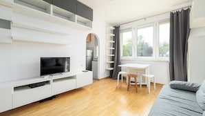 A modern living room with a flat-screen TV, white furniture, a dining area, and a large window with curtains. - RentPlanet - Studio Anielewicza (Warsaw)