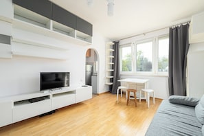 A modern living room with a flat-screen TV, white furniture, a dining area, and a large window with curtains. - RentPlanet - Studio Anielewicza (Warsaw)
