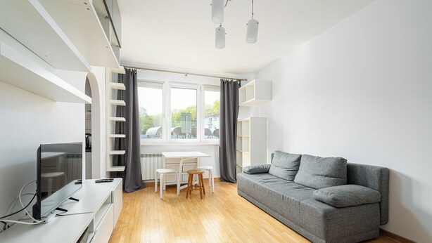 A modern living room with a grey sofa, a TV, a white table with chairs, and a window with curtains. - RentPlanet - Studio Anielewicza (Warsaw)
