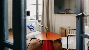 Una habitación con un sofá blanco, una mesa roja, un televisor montado en la pared y una ventana con cortinas.