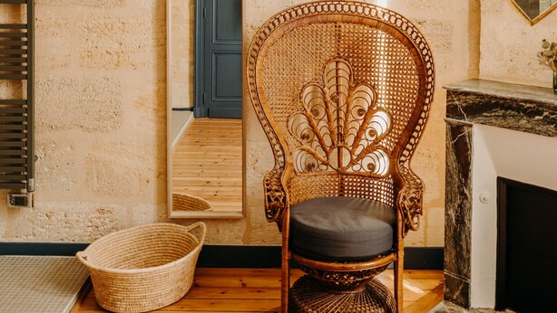 Una habitación con pared de ladrillo rústico, una silla de mimbre, suelo de madera, una lámpara blanca y un espejo que refleja una puerta azul.