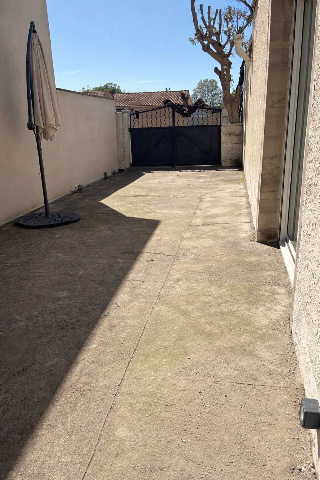 A paved patio area with a metal gate, a concrete wall, and a tree.