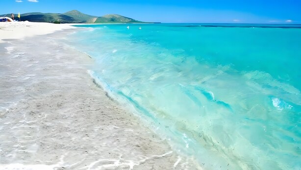 Uma praia com águas turquesa cristalinas, areia branca e montanhas ao longe.