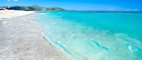 A beach with clear turquoise water, white sand, and distant mountains.