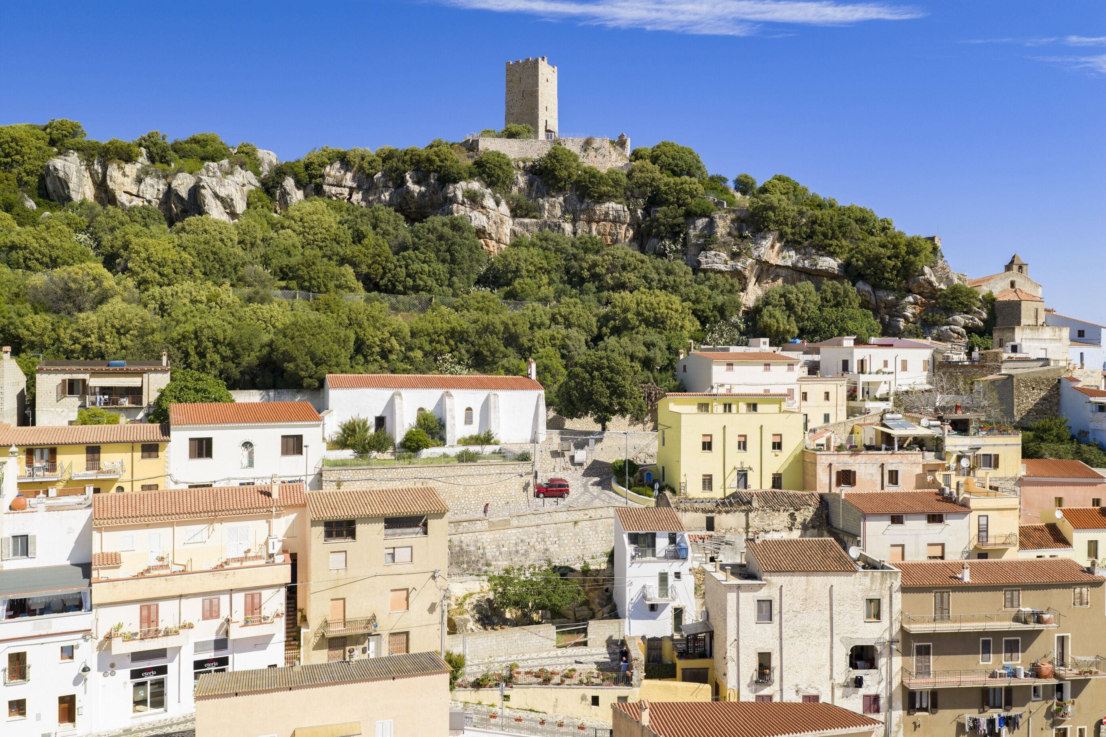 Un pueblo costero con un castillo imponente en una colina, edificios con tejados de teja anaranjada y una calle estrecha.
