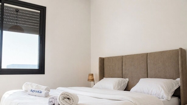 A neatly made bed with white linens and rolled towels, a headboard, a bedside lamp, and a window with blinds. - Bright, refined apartment in La Marsa (Marsa)