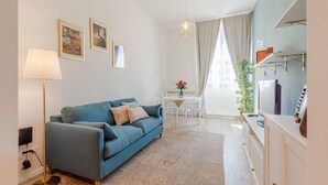 A living room with a blue sofa, a lamp, a dining table with chairs, a hanging light fixture, and a kitchen area with cabinets and a refrigerator. - Le Case di Sonia Maison Mami' (Fucecchio)