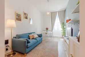 Ein Wohnzimmer mit blauem Sofa, einer Lampe, einem Esstisch mit Stühlen, einer Pendelleuchte und einer Küchenzeile mit Schränken und einem Kühlschrank.