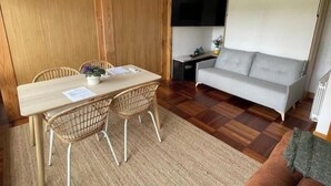 Ein Zimmer mit einem Holzesstisch, zwei Korbstühlen, einem grauen Sofa, einem kleinen Schreibtisch mit Stuhl und einem an der Wand befestigten Fernseher.