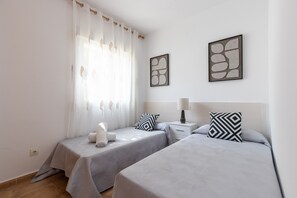 A hotel room with two beds, a window with sheer curtains, a bedside table with a lamp, and abstract wall art.