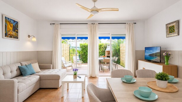 A modern living room with a sofa, coffee table, and dining area, connected to a balcony with outdoor seating.