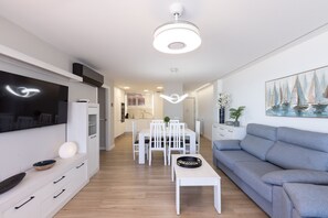 A modern living room with a flat-screen TV, a dining area with white furniture, a grey sofa, and a ceiling light fixture. - Mas d'En Gran Apartment - Vilafortuny Beach (Cambrils)