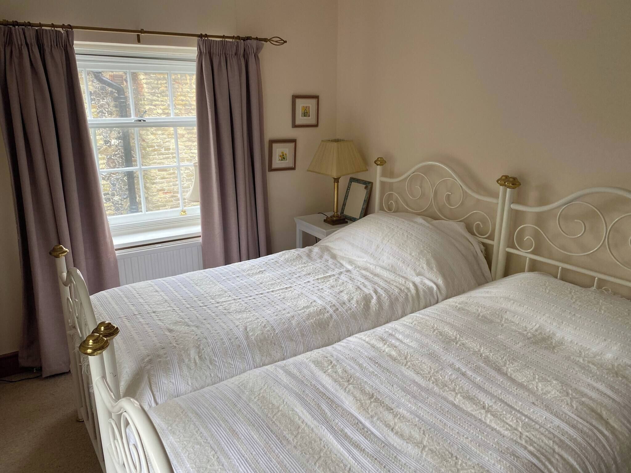 A bedroom with two beds, a window with curtains, a bedside table with a lamp, and framed pictures on the wall.