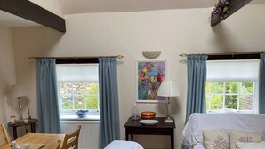 A room with a wooden dining table, two chairs, a bed with white bedding, a small bedside table with a lamp, a painting on the wall, and a window with blue curtains. - Kent Apartment 2 bed in Sandwich, With Free Parking (Sandwich)