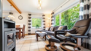 Ein Wohnzimmer mit einer Couch, einem Liegesessel, einem Esstisch mit Stühlen, einem Kamin und großen Fenstern mit Vorhängen.