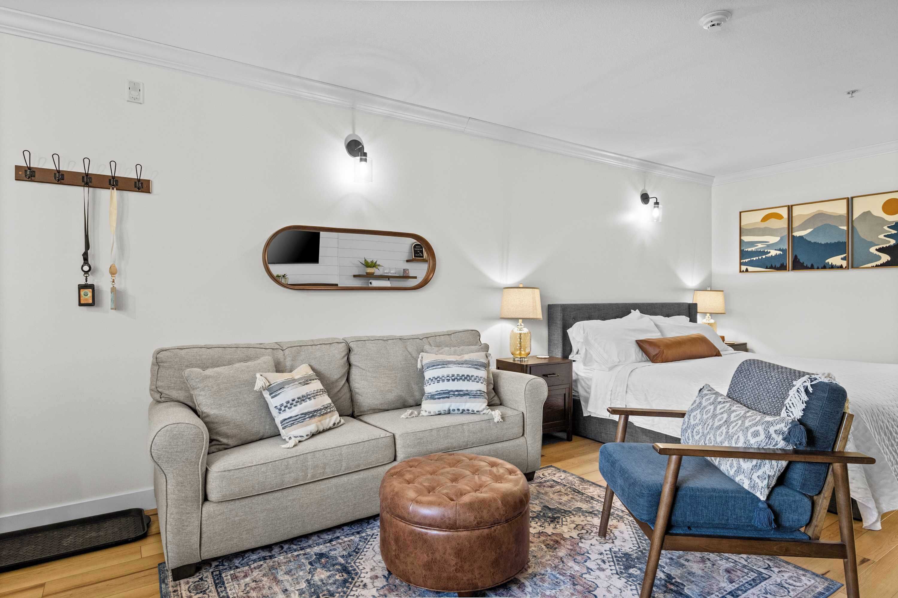 A modern room with a sofa, ottoman, chair, bed, bedside tables, lamps, wall art, and a coat rack.